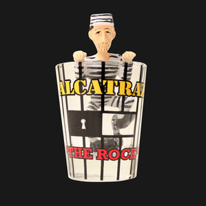 A shot glass with "Alcatraz The Rock" design printed on the outside. A resin figure of a prisoner is on the inside of the shot glass climbing out of the glass